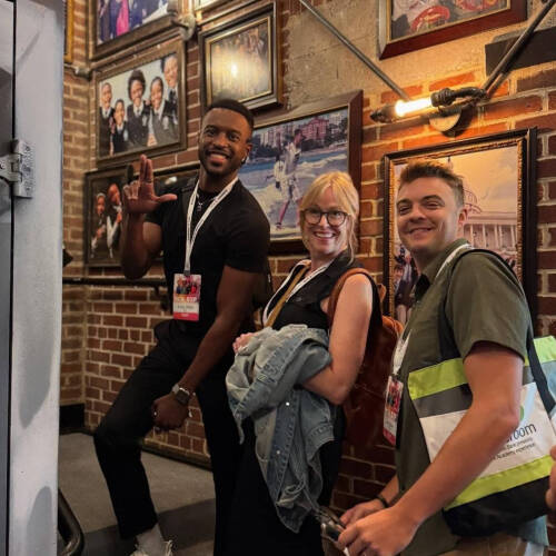 Three people pose together in a hallway lined with framed photos on a brick wall, wearing lanyards from the "RCA Exp